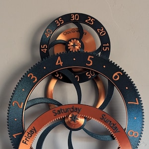 May include: A mechanical wall clock featuring interlocking gears in black and copper tones. The design includes circular rings with printed numbers and days of the week, specifically Monday through Sunday, and numerical markings for timekeeping.