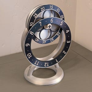 Könnte beinhalten: Ein silberfarbener mechanischer Timer mit zwei rotierenden, nummerierten Zifferblättern. Die Zifferblätter haben schwarze Zahlen und Markierungen auf weißem Hintergrund. Der Timer ist auf einem kreisförmigen Sockel montiert und steht aufrecht.