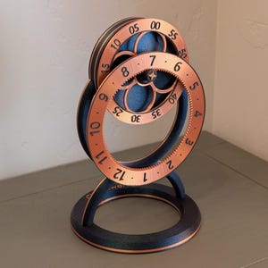 Puede incluir: Un reloj decorativo con un diseño de engranaje único. Presenta dos esferas circulares de color cobre con números y un detalle central en forma de estrella. La base y la estructura de soporte son negras.