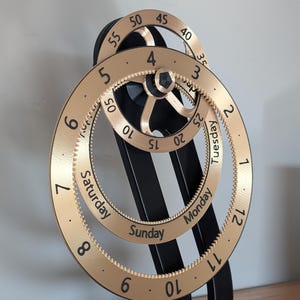 May include: A mechanical calendar clock featuring a black stand and two rotating gold rings. The outer ring displays numbers 1 through 12 and days of the week, while the inner ring shows numbers 5 through 55 in increments of five and 1 through 5.