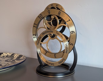 May include: A mechanical desk clock featuring a gold-colored circular frame with black numerals 5 through 12 and a smaller inner gold ring displaying the days of the week from Monday to Sunday. The structure is supported by a black circular base.
