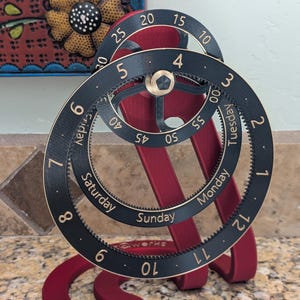 May include: A mechanical calendar clock with a red stand and black circular rings. The rings feature gold numbers for hours and minutes, and the days of the week including Monday, Tuesday, Friday, Saturday, and Sunday. The base is marked with the text qwerks.