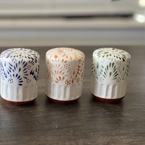 Puede incluir: Tres saleros cilíndricos de cerámica con base blanca y fondo color terracota. Cada salero presenta un patrón floral pintado a mano en azul, naranja o verde, con estrías verticales en la parte inferior y pequeños orificios en la parte superior.