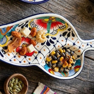 Puede incluir: Una fuente de cerámica con forma de hoja, pintada a mano con diseños florales y geométricos coloridos. Contiene diversos aperitivos, como frutos secos y galletas saladas. También se ven otros platos y cuencos de cerámica.