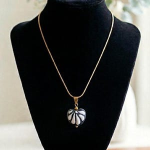 Puede incluir: Un collar de cadena dorada con un colgante en forma de corazón. El colgante es blanco y negro con un diseño floral. El collar se exhibe sobre un busto de terciopelo negro.
