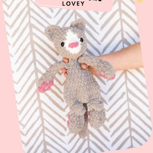 May include: A crochet cat lovey named Kai the Cat with a grey textured body, a white patch on its face, black eyes, and a pink nose. The plush toy features pink accents on the paws and feet, crafted from soft yarn with a visible knit pattern.