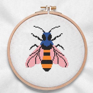 May include: A cross-stitch embroidery of a bee in a wooden hoop. The bee features a blue head, pink wings, and a black and orange striped body. The embroidery is on white fabric.