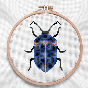 May include: A cross-stitch embroidery of a blue beetle with black spots and orange accents, set in a wooden embroidery hoop. The beetle design is centered on a white fabric background, showcasing the detailed needlework.
