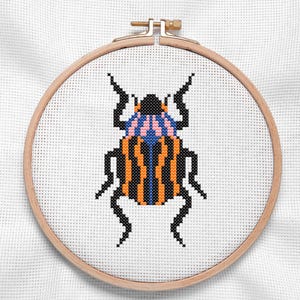 May include: A cross-stitch embroidery of a colorful beetle, featuring orange, black, blue, and pink details. The insect is centered on white fabric, framed by a wooden embroidery hoop. The design showcases a detailed, handcrafted insect.