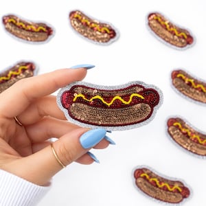 May include: A sequined hot dog patch with a silver glitter border. The hot dog is brown with red and yellow accents, and is held by a hand with blue nails. Several other patches are scattered around the main patch.