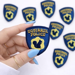 May include: A collection of blue and yellow embroidered patches with the text "SQUIRREL POLICE" and a squirrel silhouette. The patches are shield-shaped and have a yellow border. The patches are on a white background.