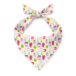 May include: White dog bandana with a colorful pattern of lobsters, jars, lemons, and other food items. The bandana has a striped background and is tied at the top. The bandana is made of a soft material and is designed for pets.