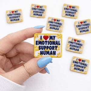 May include: Square embroidered patch with the text "I ❤️ MY EMOTIONAL SUPPORT HUMAN" in navy blue, a red heart, and a yellow floral border. Several patches are visible.