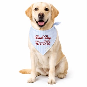 May include: A golden Labrador wearing a light blue and white striped bandana with the text "Bad Day to be a Hotdog" in red script. The dog is sitting, and the background is white.