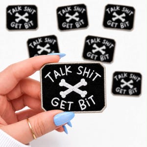 May include: Black rectangular patch with white text and a crossed bone design. The text reads "TALK SHIT GET BIT". Several patches are visible, some in the background. The patch is held by a hand with blue nails.