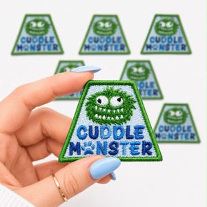 May include: Embroidered patch with a green monster face and the words "Cuddle Monster" in blue. The patch has a light blue background and a green border. Several similar patches are visible.