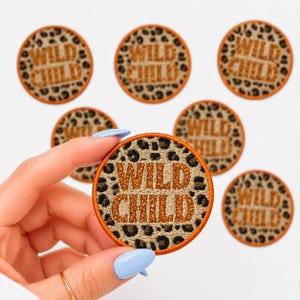 May include: Round patches with a leopard print background and the words "WILD CHILD" in glittery orange letters. The patches have an orange border. One patch is held by a hand with blue nail polish.