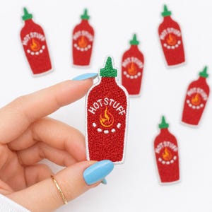 May include: Red embroidered patches in the shape of hot sauce bottles with the text "HOT STUFF" and a flame graphic. The patches have green caps and white details. One patch is held by a hand with a gold ring and blue nail polish.