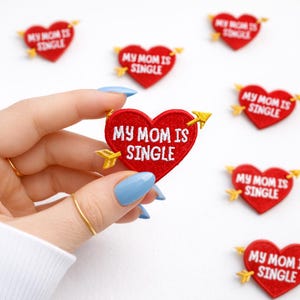 May include: Red heart-shaped patches with the text "MY MOM IS SINGLE" embroidered in white. Each patch has a yellow arrow design. The patches are scattered on a white background.