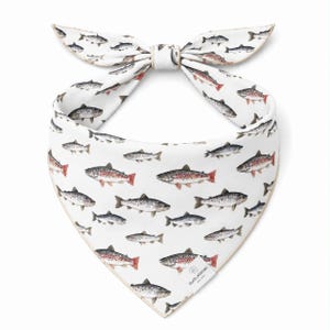 May include: White dog bandana with a repeating pattern of fish in shades of gray, red, and brown. The bandana is tied in a bow at the top. The bottom corner has a small logo.
