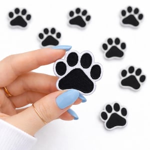 May include: Black and white paw print patches. The patches feature a black paw print design with a white border. Several patches are scattered on a white background. One patch is held up by a hand with blue nail polish.