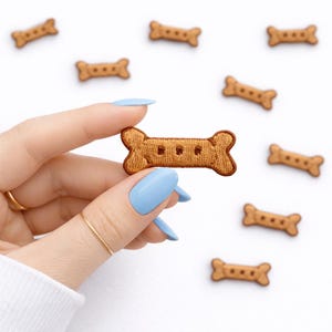 May include: A collection of brown, bone-shaped embroidered patches with three small holes in the center. The patches are scattered on a white background, with one patch being held up by a hand with blue nail polish.
