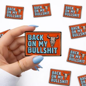 May include: Orange embroidered patch with the text "BACK ON MY BULLSHIT" in light blue, featuring a bull skull graphic. Several patches are visible. The patches are rectangular.