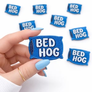 May include: Blue embroidered patch in the shape of a pillow with the words "BED HOG" in white. Several patches are scattered on a white background. The patch is held by a hand with blue nail polish.