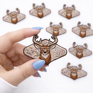 May include: Embroidered deer patches with brown and tan details. The patches feature a deer head with large antlers, set against a diamond-shaped background with floral patterns. Several patches are visible, with one held up by a hand.