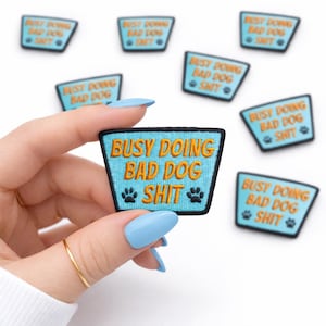 May include: Embroidered patch with the text "BUSY DOING BAD DOG SHIT" in orange thread on a turquoise background. The patch includes two paw prints and a black border. Several similar patches are scattered around.