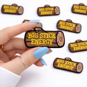 May include: Embroidered patch in the shape of a log with the text "BIG STICK ENERGY" in yellow. The patch is brown with yellow stitching. Several patches are visible on a white background.
