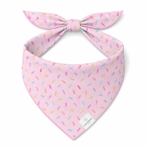 May include: Pink dog bandana with a bow, patterned with colorful sprinkles. The bandana is made of soft fabric and features a small white tag with the brand name. A stylish accessory for pets.