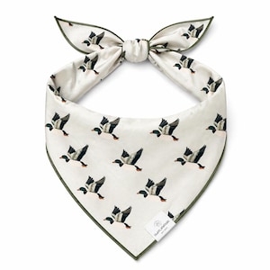May include: Cream-colored bandana with a repeating pattern of flying ducks. The bandana has a dark green trim and is tied at the top. A small white tag with text is visible on the bottom right corner. The bandana is likely made of a soft fabric.