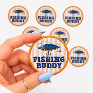 May include: Embroidered patch with a fishing lure design. The patch features a blue and orange fish, the words "FISHING BUDDY", and a striped background. Several patches are visible.