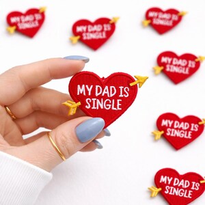 May include: Red heart-shaped patches with the text "MY DAD IS SINGLE" embroidered in white. Each patch has a yellow arrow through it. Several patches are scattered on a white surface.