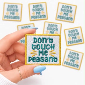 May include: Square embroidered patch with the text "Don't Touch Me Peasant" in teal and yellow on a white background. The patch has a gold border. Several similar patches are visible.