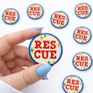 May include: Embroidered patches with the word "RES CUE" in red, surrounded by a blue border and star accents. The patches are circular and have a cream-colored background. Several patches are scattered around the main patch.