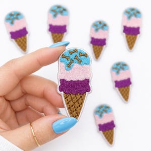 May include: Embroidered ice cream cone patches in pink, purple, and blue with bone-shaped sprinkles. The patches have a brown cone and are held by a hand with a gold ring and blue nail polish. Several other patches are scattered in the background.