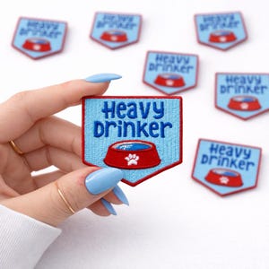 May include: Embroidered patches with a light blue background and a red border. The patches feature the words "Heavy Drinker" above a red dog bowl with a white paw print. Several patches are visible.