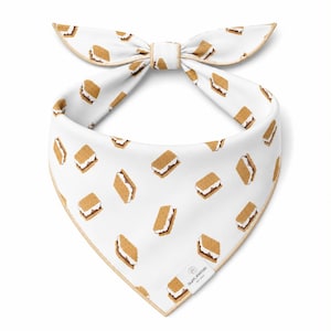 May include: White pet bandana with a repeating pattern of s'mores. The bandana has a bow tie design and is trimmed with a light brown border. The s'mores are brown, white, and tan.