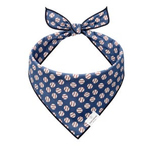 May include: Blue dog bandana with a baseball pattern. The bandana features a repeating pattern of white baseballs with red stitching on a blue background. The edges are trimmed with black. A small white tag with text is attached.