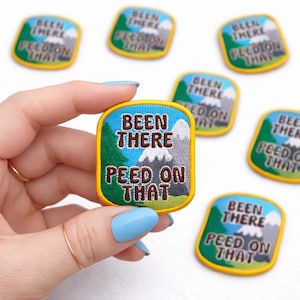 May include: Embroidered patch with the text "BEEN THERE PEED ON THAT" in brown lettering. The patch features a mountain and tree design with a blue sky background and yellow border. Several similar patches are visible.