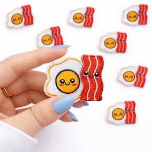 May include: Embroidered patches featuring a smiling fried egg and strips of bacon. The egg has a white border and a yellow yolk with a happy face. The bacon is red with white stitching and also has a smiling face. Several patches are scattered on a white background.