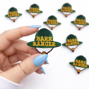May include: Embroidered patch featuring a green ranger hat with the words "BARK RANGER" in yellow. The patch has a white and brown brim and is surrounded by other similar patches. The patches are likely for pet owners.