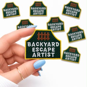May include: Embroidered patch with the text "BACKYARD ESCAPE ARTIST" in white on a dark green background. The patch has a yellow border and a brown fence design at the top. Several patches are visible.