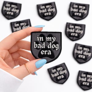 May include: A collection of black and gray patches with the phrase "in my bad dog era" embroidered in white. One patch is held by a hand with a gold ring and light blue nails. The patches feature a paw print design.