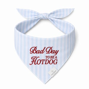 May include: A light blue and white striped dog bandana with a bow. The bandana features the embroidered text "Bad Day to be a Hotdog" in red. A small white tag with text is attached to the bottom.