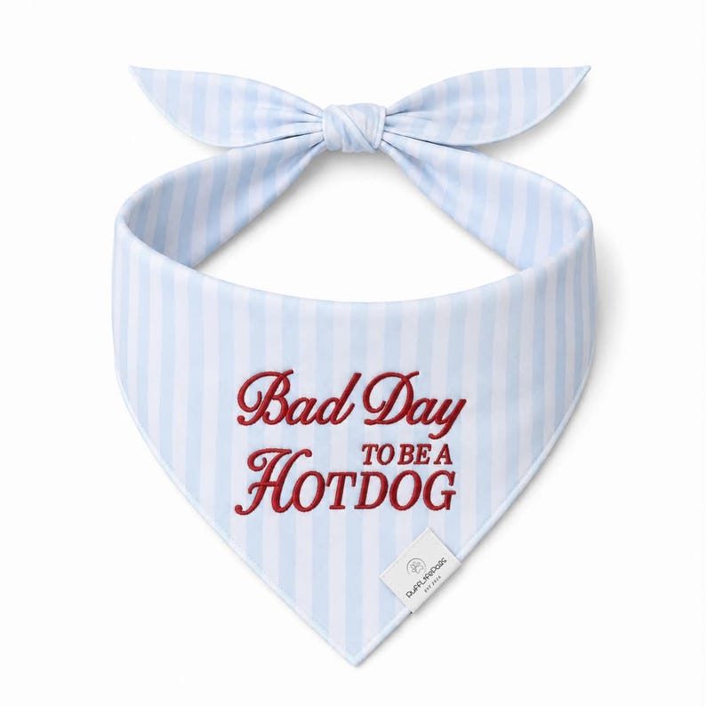 May include: A light blue and white striped dog bandana with a bow. The bandana features the embroidered text "Bad Day to be a Hotdog" in red. A small white tag with text is attached to the bottom.