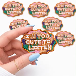 May include: Embroidered patch with the text "I'M TOO CUTE TO LISTEN" in white letters. The patch has a colorful design with pink, green, purple, and brown accents. Several patches are visible.