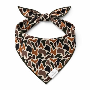 Puede incluir: Un pa&ntilde;uelo con estampado de camuflaje con un borde verde oscuro y un lazo en la parte superior. El pa&ntilde;uelo presenta un patr&oacute;n repetido de formas marrones, negras y verdes sobre un fondo crema. Una peque&ntilde;a etiqueta blanca con texto est&aacute; adjunta en la parte inferior.